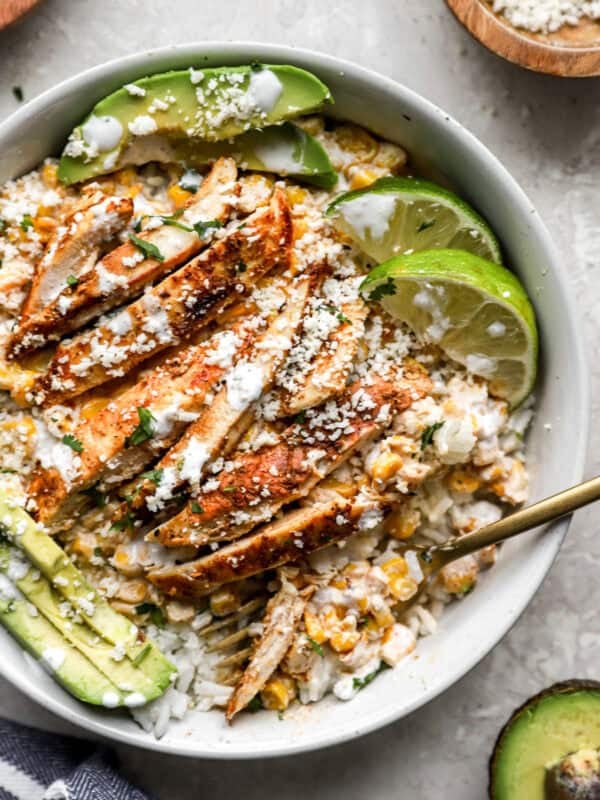 Street corn chicken bowl with sliced grilled chicken, creamy corn mixture, white rice, avocado slices, cotija cheese, and lime wedges.