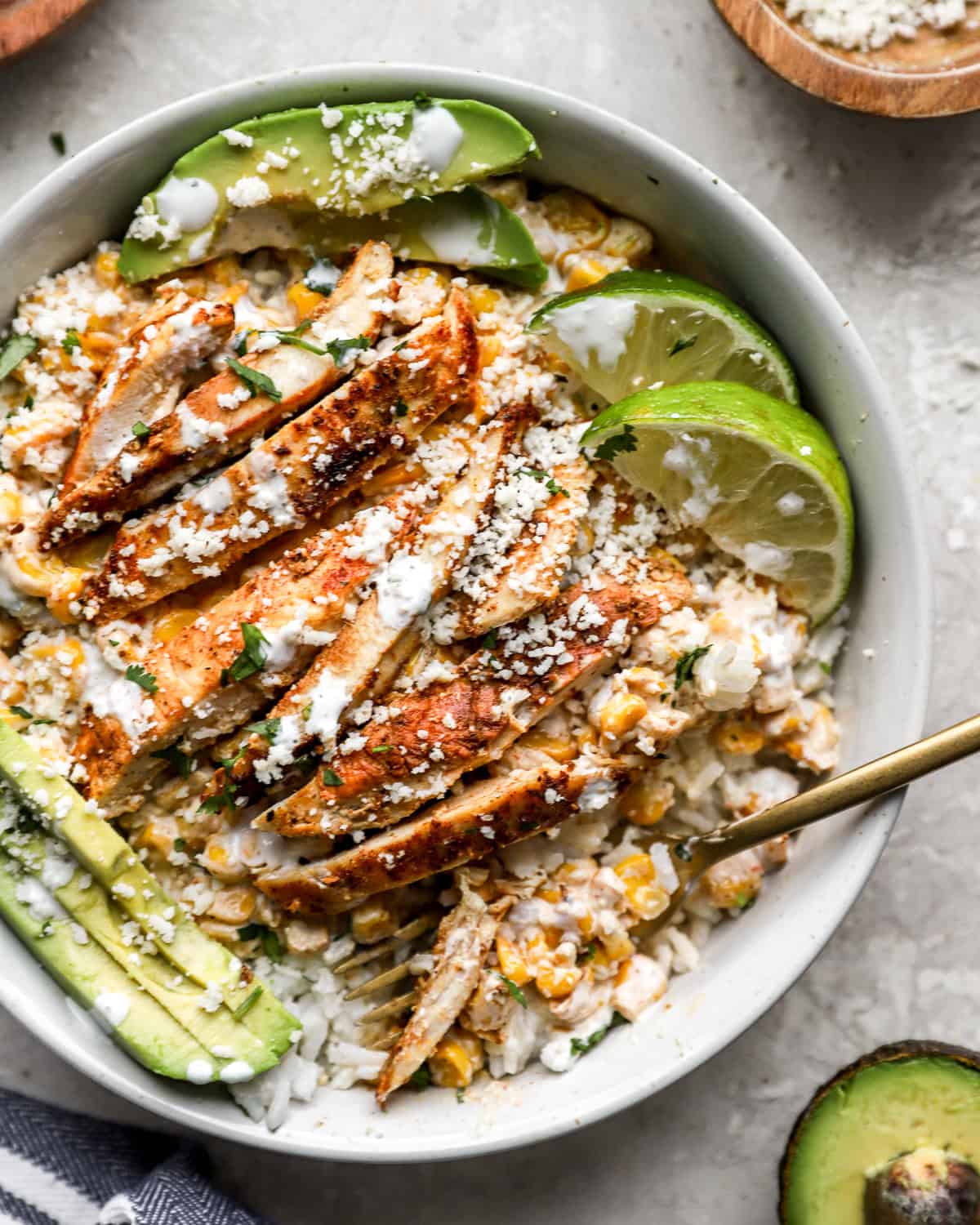Street corn chicken bowl with sliced grilled chicken, creamy corn mixture, white rice, avocado slices, cotija cheese, and lime wedges.