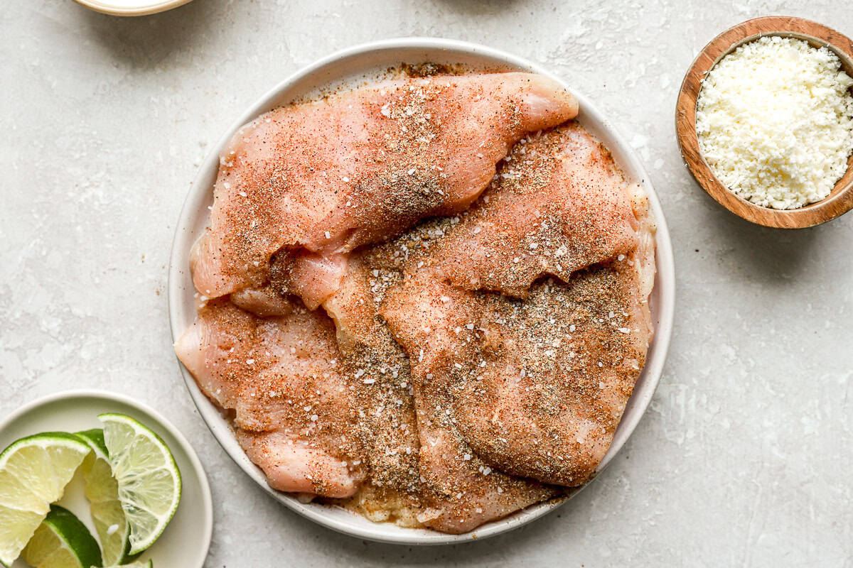 Raw chicken breasts seasoned with spices on a plate with lime wedges and crumbled cotija cheese nearby.
