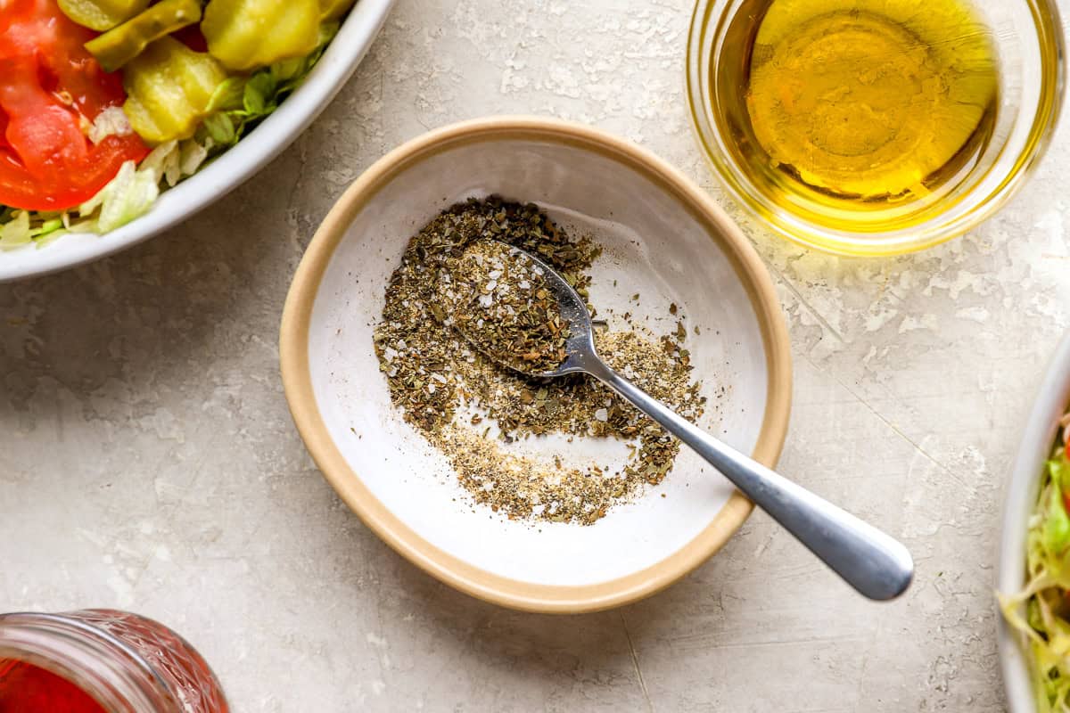 Small bowl of oregano, garlic powder, salt, and pepper seasoning mix with spoon next to bowls of sub salad