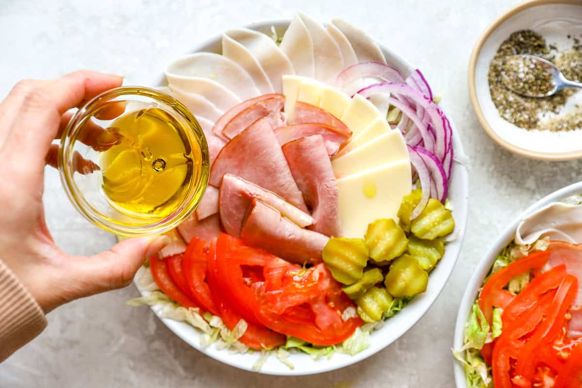 Hand drizzling olive oil over an Italian sub salad bowl with deli meats, cheese, tomatoes, and pickles