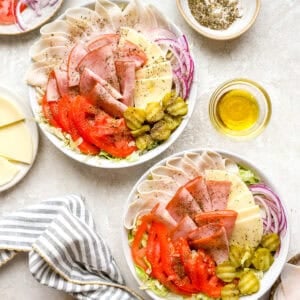 Two bowls of Italian sub salad with turkey, ham, provolone, tomatoes, pickles, and red onion over shredded lettuce with oil and seasoning on the side