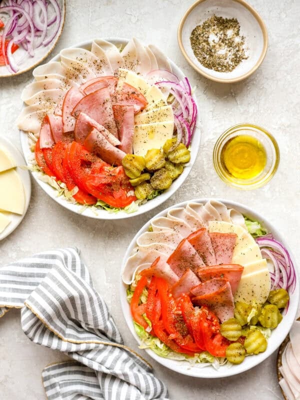 Two bowls of Italian sub salad with turkey, ham, provolone, tomatoes, pickles, and red onion over shredded lettuce with oil and seasoning on the side