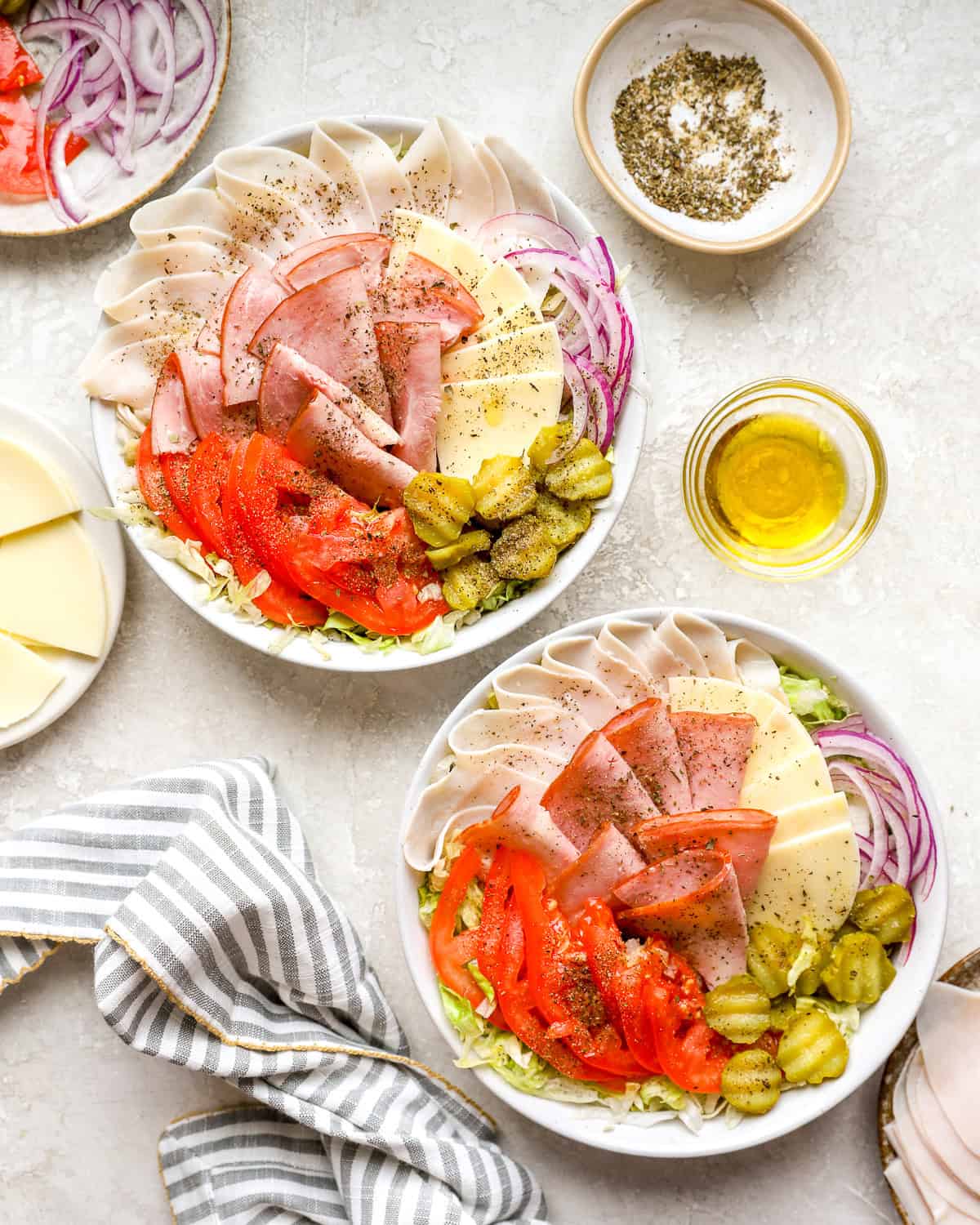 Two bowls of Italian sub salad with turkey, ham, provolone, tomatoes, pickles, and red onion over shredded lettuce with oil and seasoning on the side