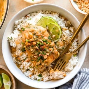Coconut chicken served over white rice topped with chopped peanuts, cilantro, and lime slices in a bowl