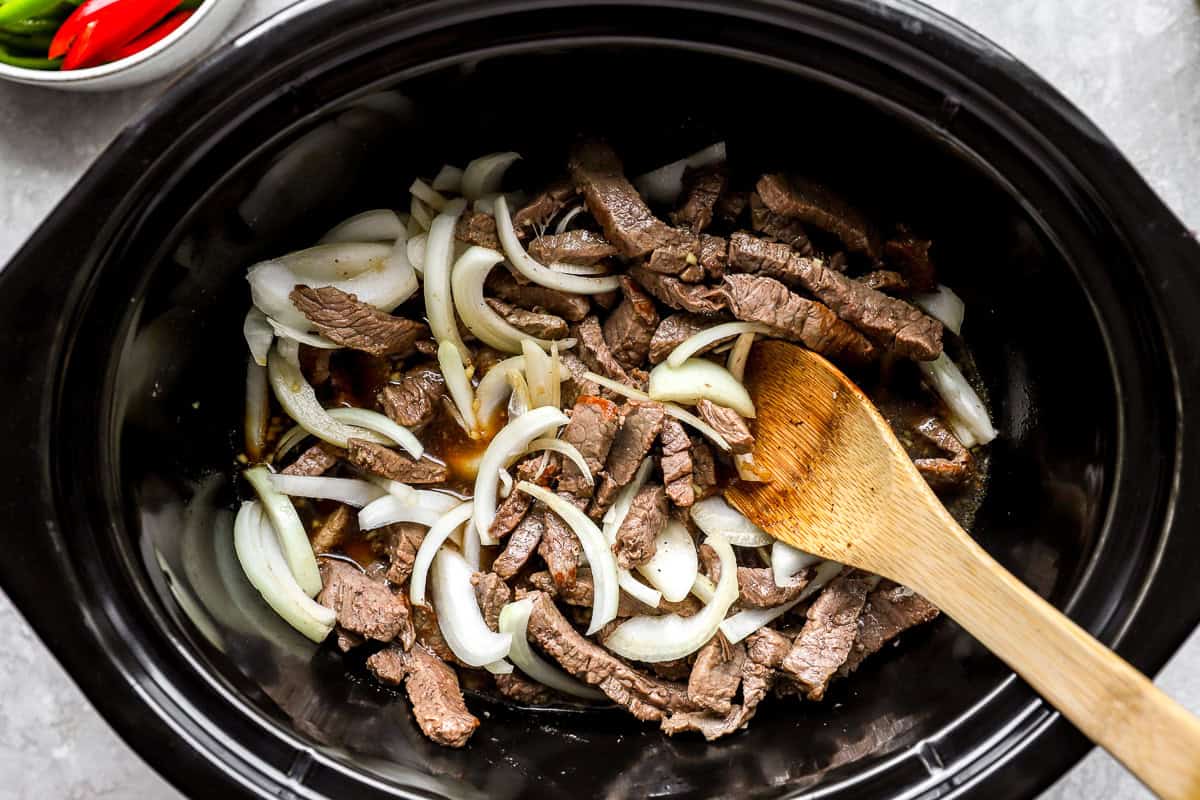 Browned steak and sliced onions added to a crockpot with pepper steak sauce.