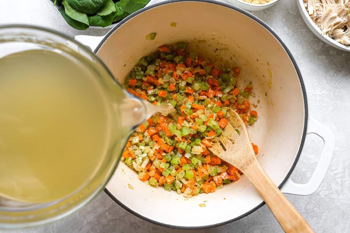 Chicken broth being poured into sautéed vegetables to make lemon chicken orzo soup