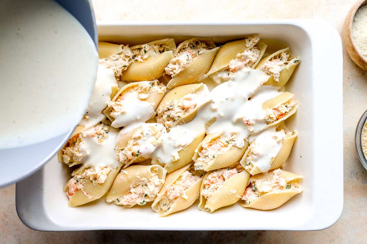 Cream sauce being poured over seafood stuffed shells before baking.