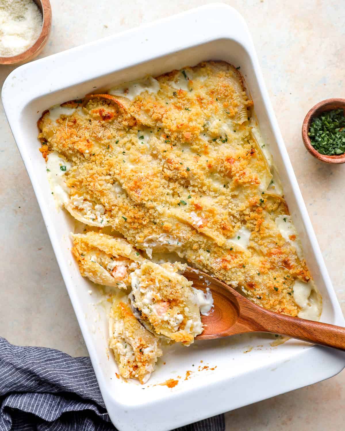 Baked seafood stuffed shells in a white casserole dish topped with golden breadcrumbs and parmesan, with a spoon scooping out a shell.