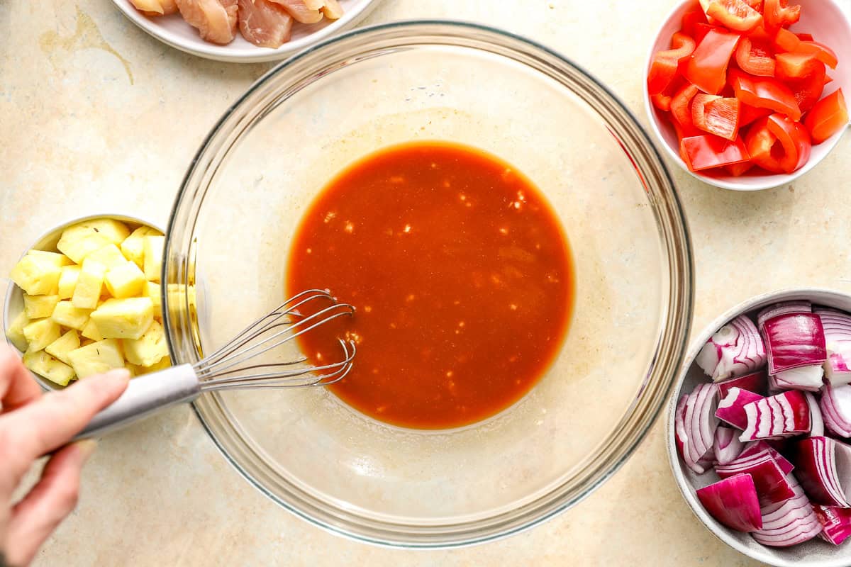Whisking Hawaiian chicken sauce in a glass bowl with pineapple, peppers, and onions nearby