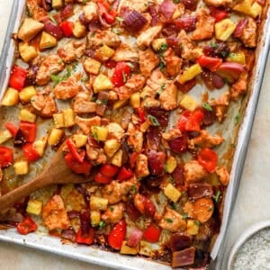 Sheet pan Hawaiian chicken with pineapple, red peppers, and red onion caramelized on a baking sheet with a wooden spoon serving.
