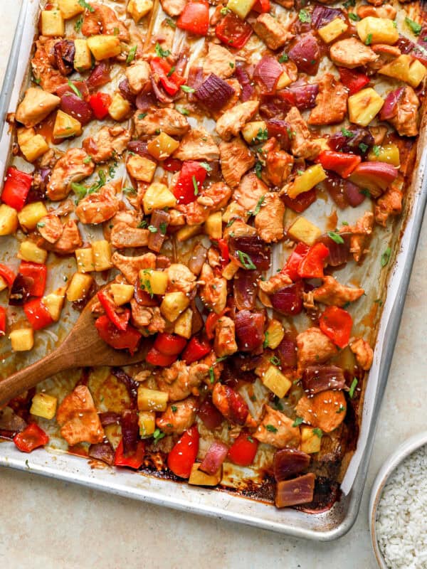 Sheet pan Hawaiian chicken with pineapple, red peppers, and red onion caramelized on a baking sheet with a wooden spoon serving.