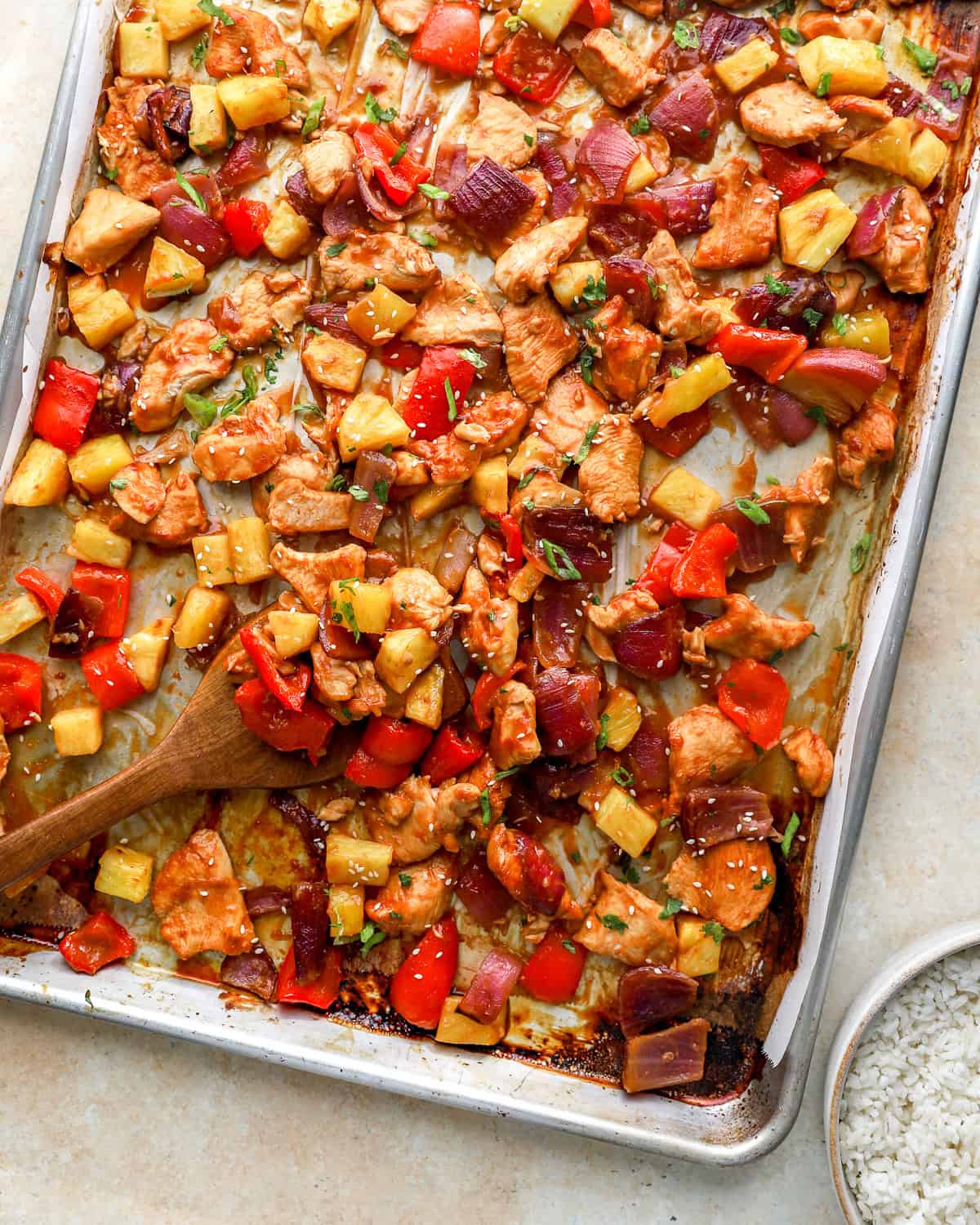 Sheet pan Hawaiian chicken with pineapple, red peppers, and red onion caramelized on a baking sheet with a wooden spoon serving.