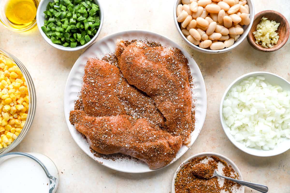 Seasoned chicken breasts with chili spices on a plate surrounded by corn, beans, onion, jalapeños, and garlic