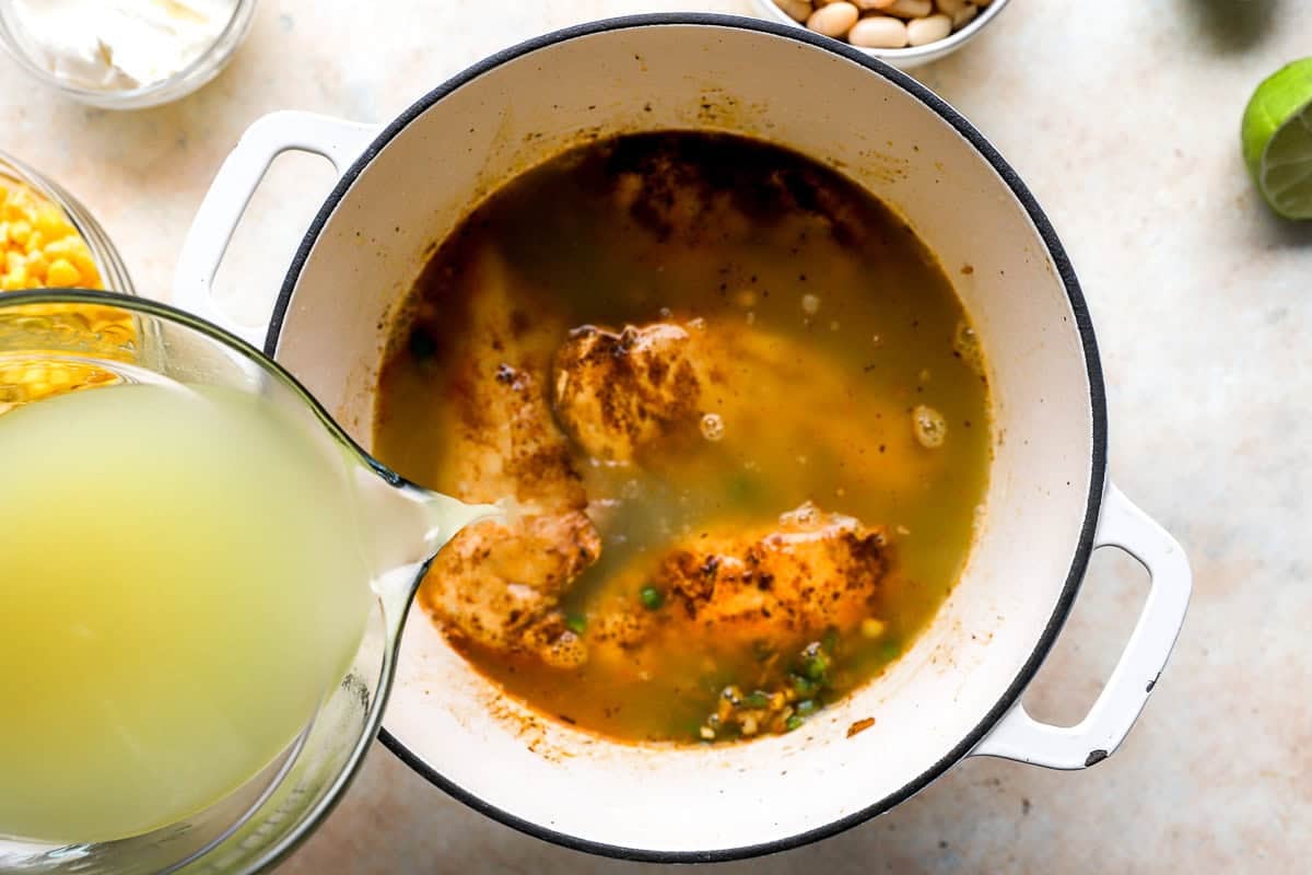 Chicken broth being poured into a Dutch oven with browned chicken and aromatics