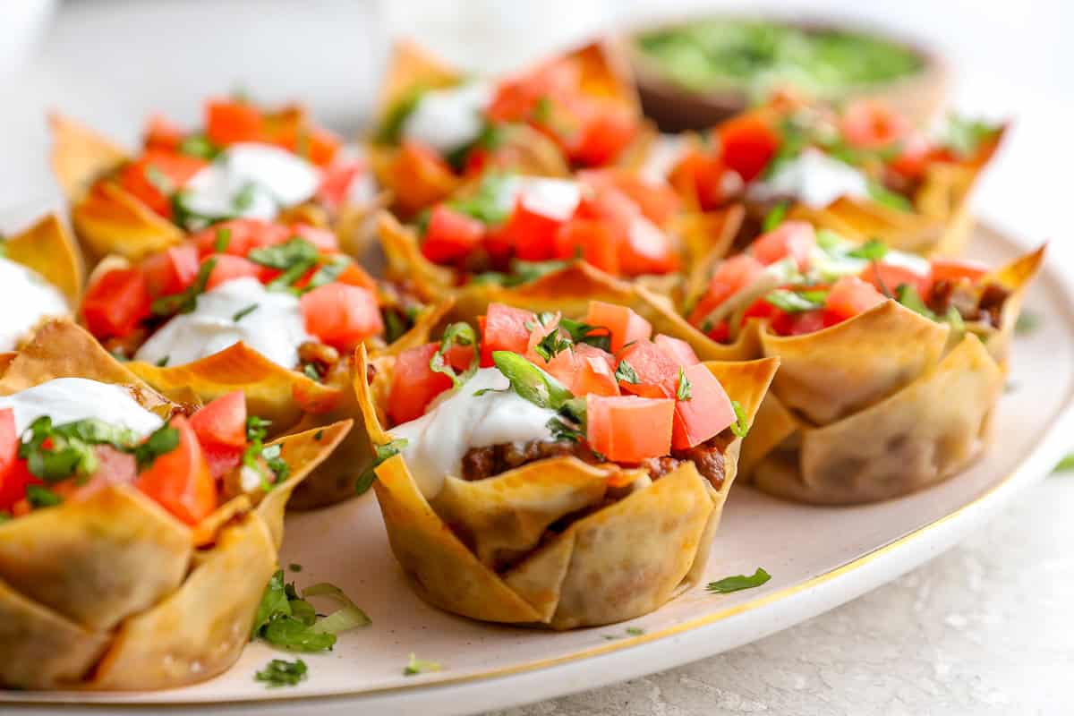 Close up of crispy taco cups made with wonton wrappers filled with taco meat and topped with sour cream, diced tomatoes, and cilantro on a platter.