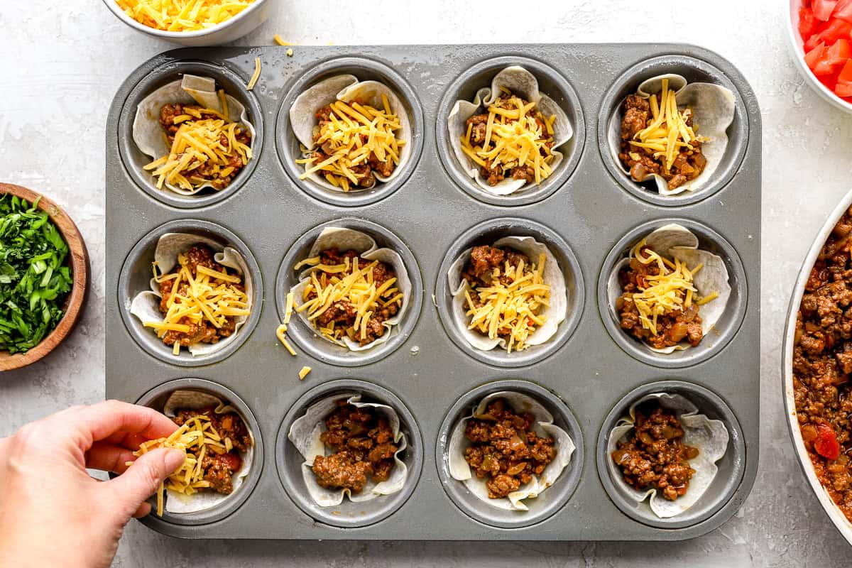 Wonton taco cups filled with taco meat and shredded cheddar cheese in a muffin pan