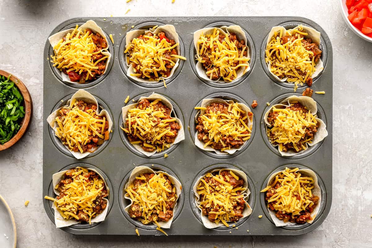 Unbaked taco cups filled with seasoned beef and shredded cheese in a muffin pan