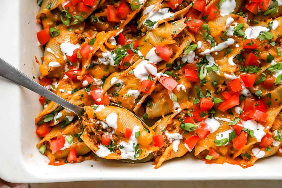 Spoon lifting taco stuffed shells from casserole dish showing cheesy taco beef filling.