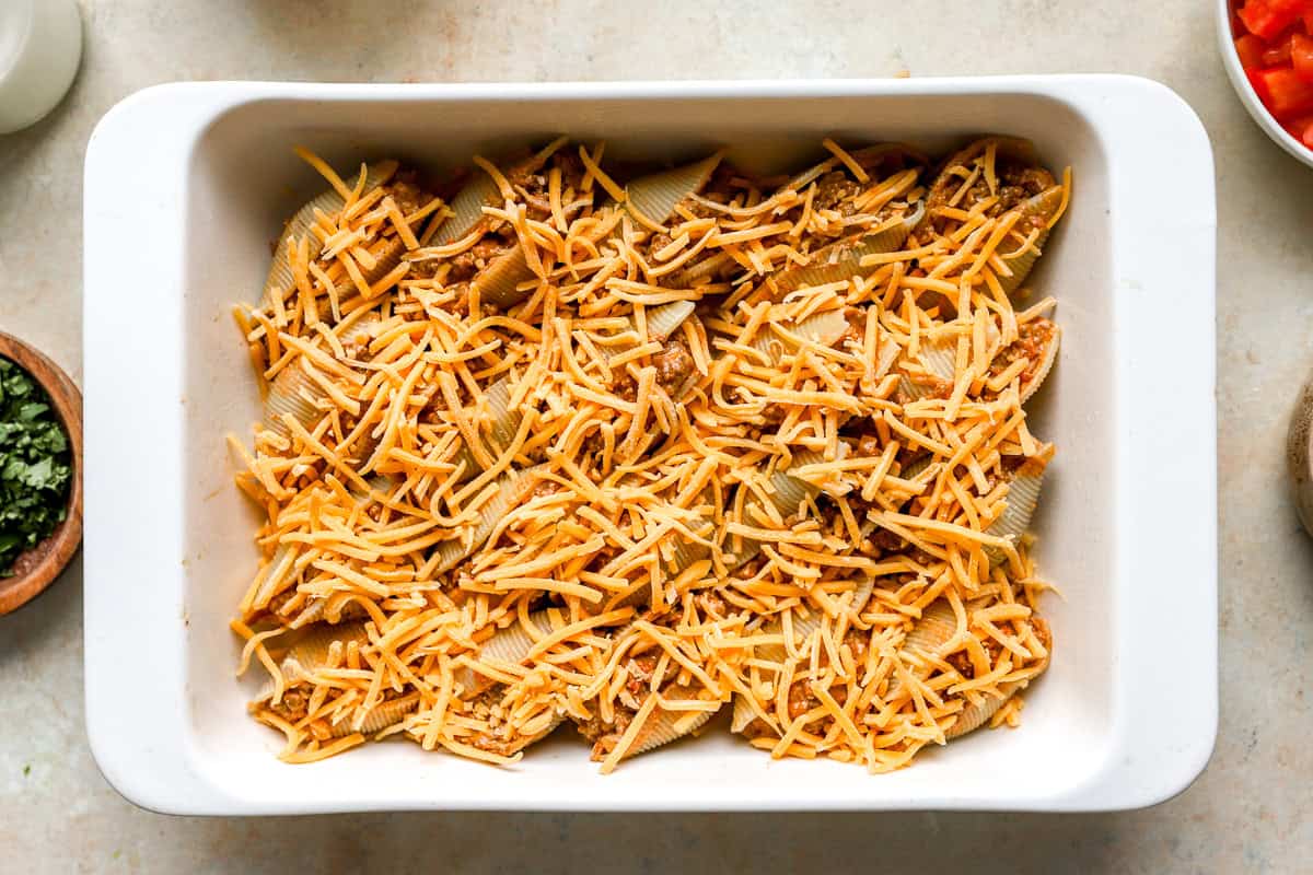 Taco stuffed shells topped with shredded cheddar cheese before baking.