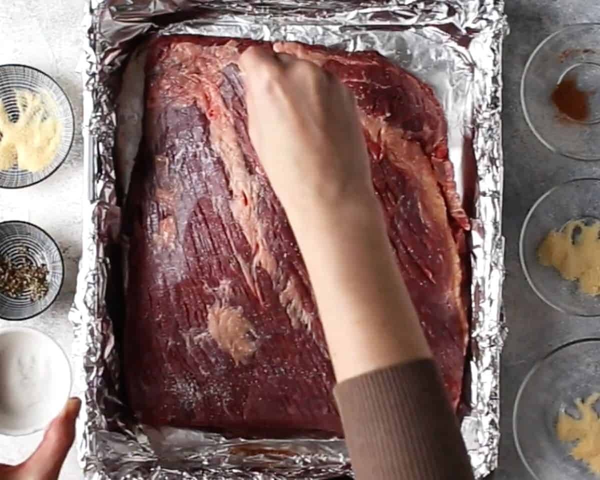 Raw beef brisket in foil-lined pan being seasoned by hand