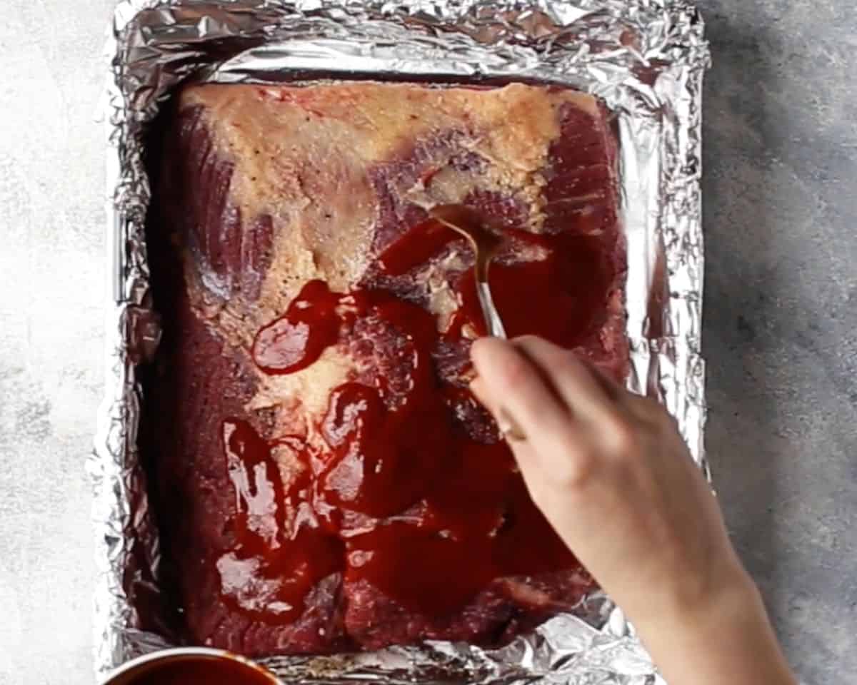 BBQ sauce being poured over seasoned brisket in baking pan