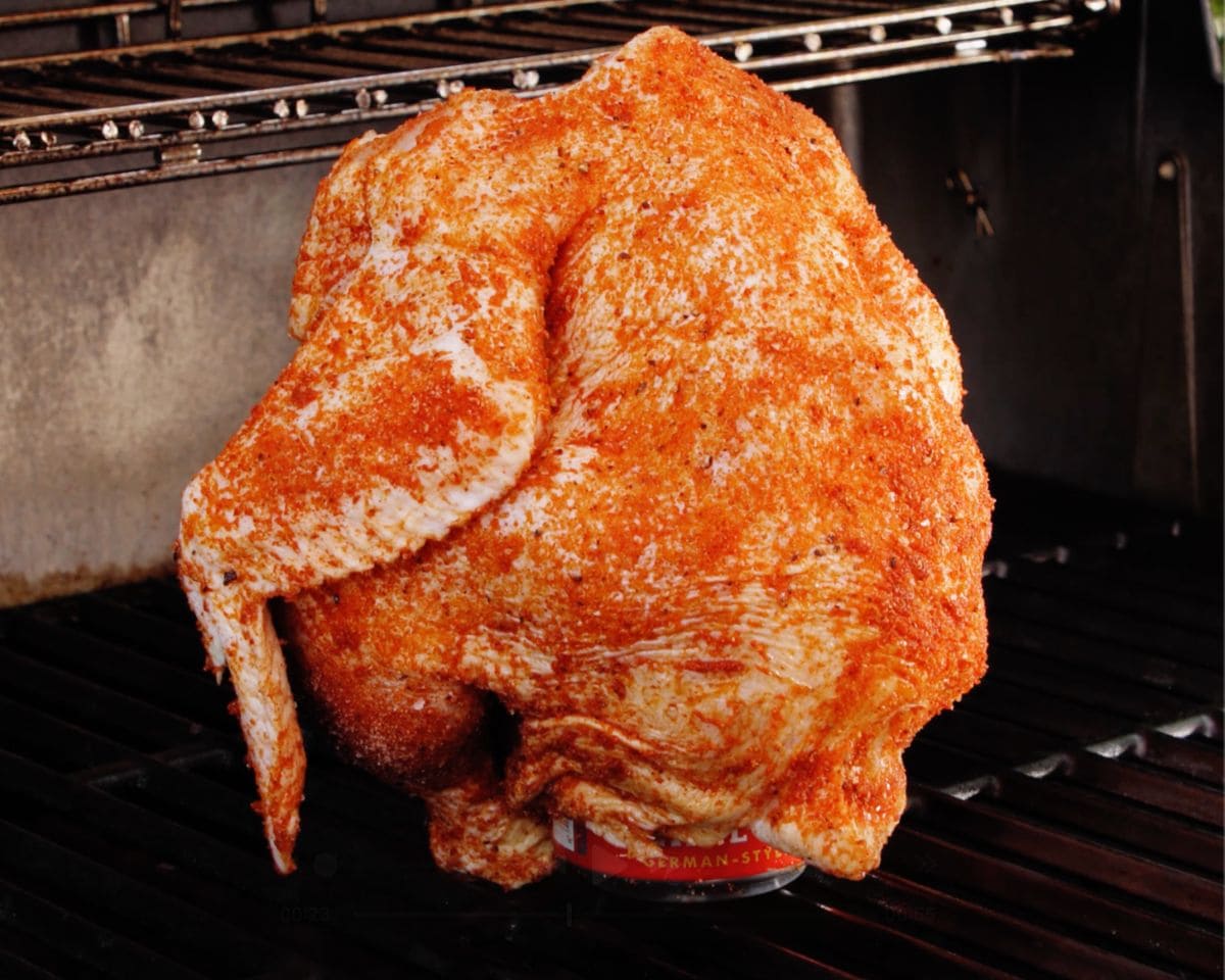 Whole chicken coated in seasoning sitting upright on a beer can on a grill, ready to cook over indirect heat.