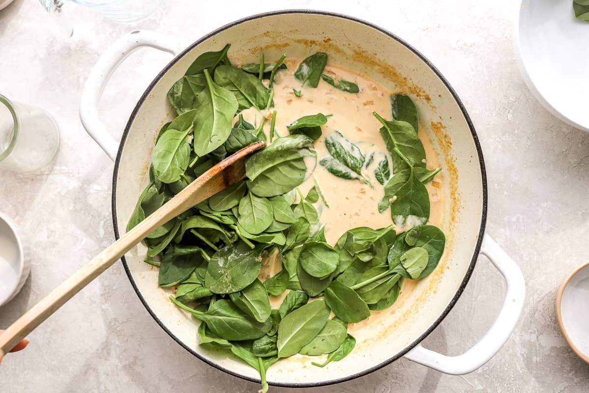 Fresh baby spinach being added to the creamy sauce in the skillet