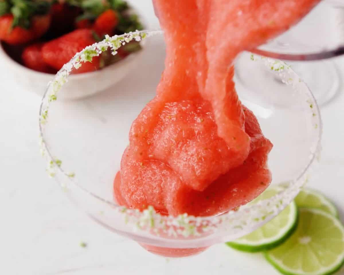 Thick frozen strawberry margarita being poured into a salt-rimmed margarita glass.