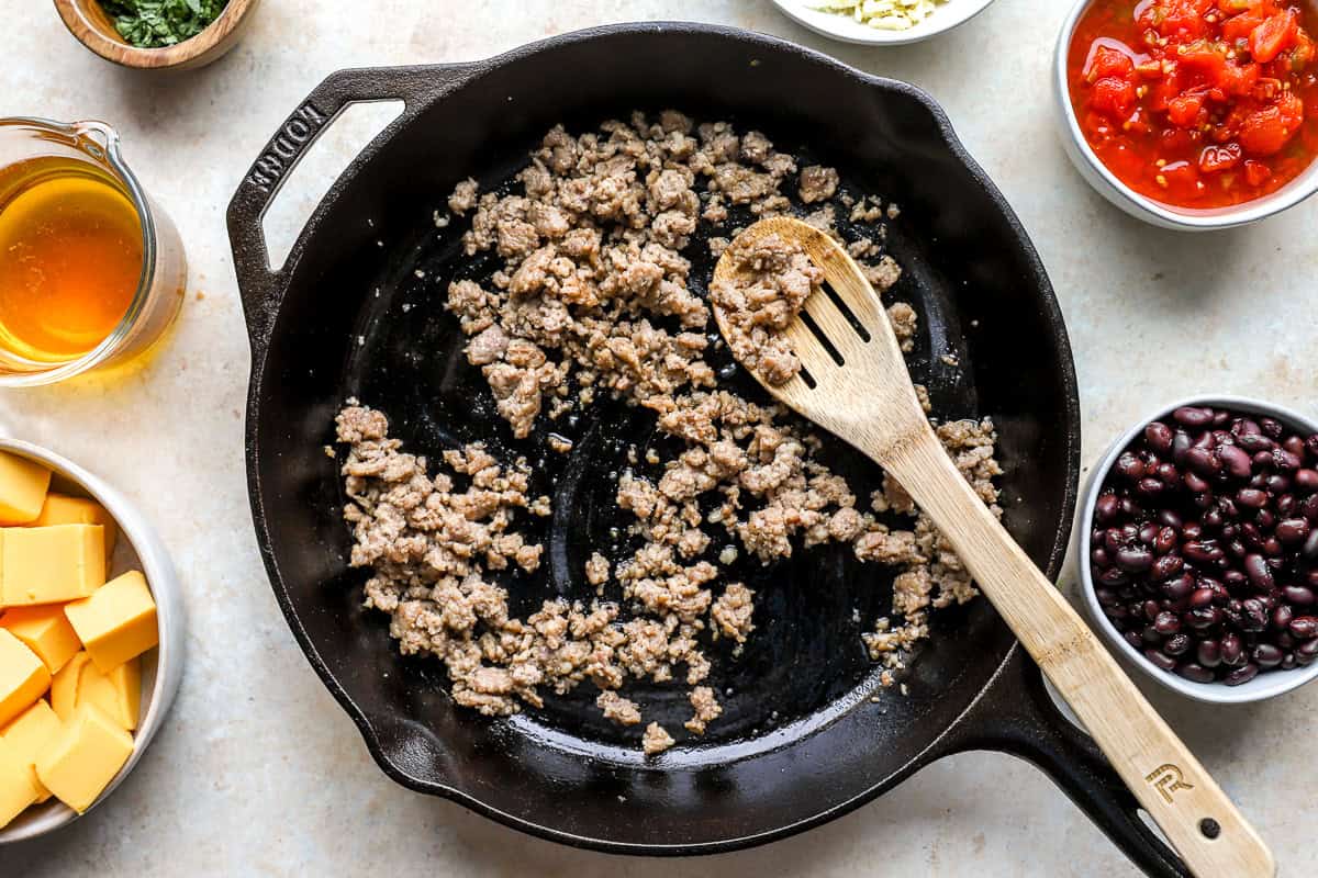 Browning pork sausage in a cast iron skillet with Velveeta cubes, beans, and Rotel ingredients nearby