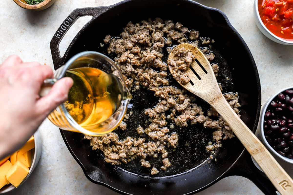 Pouring beer into browned sausage in a skillet to build flavor for loaded cowboy queso
