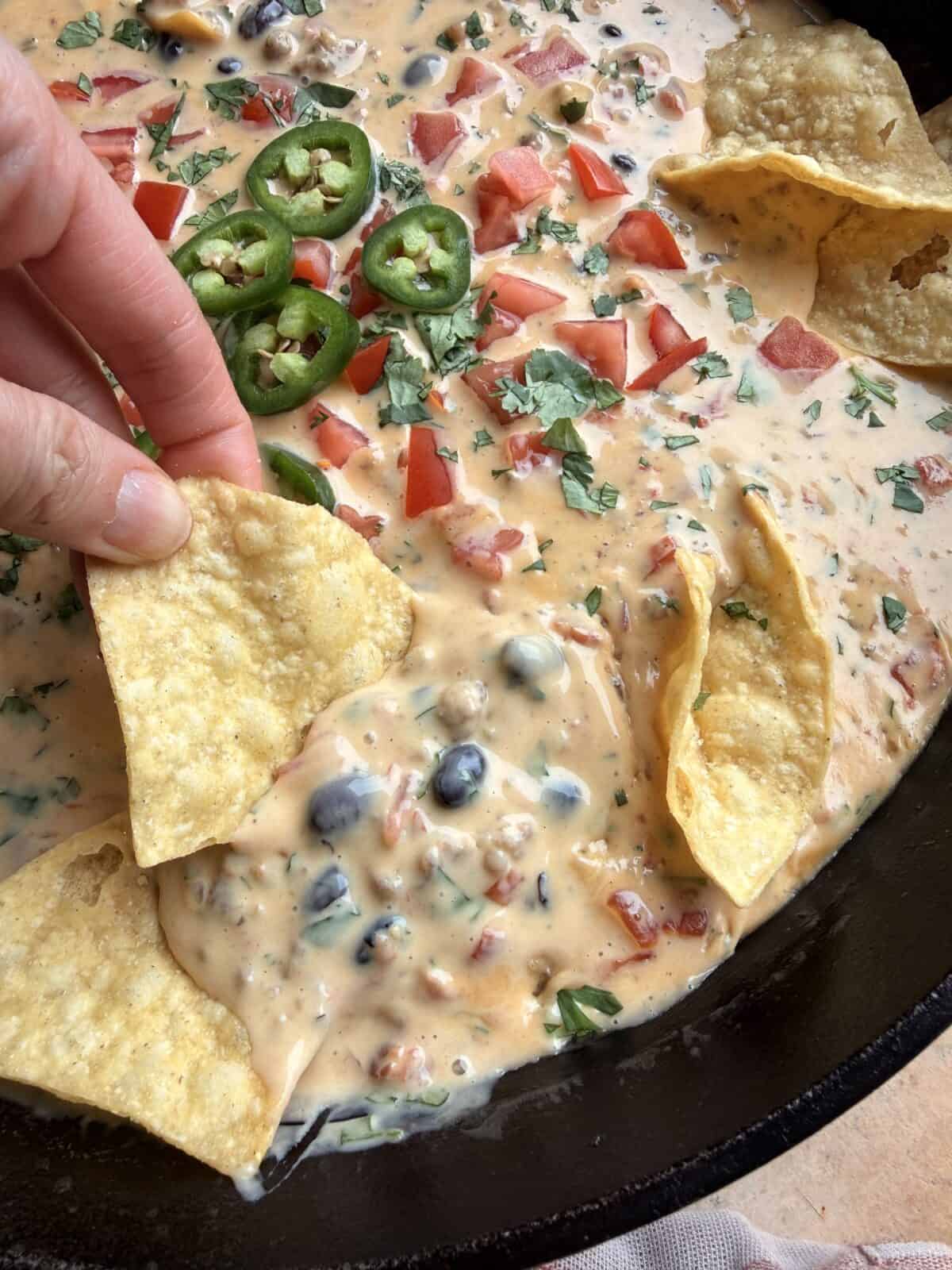 Close-up of tortilla chips dipped into creamy cowboy queso with beans, sausage, and fresh toppings