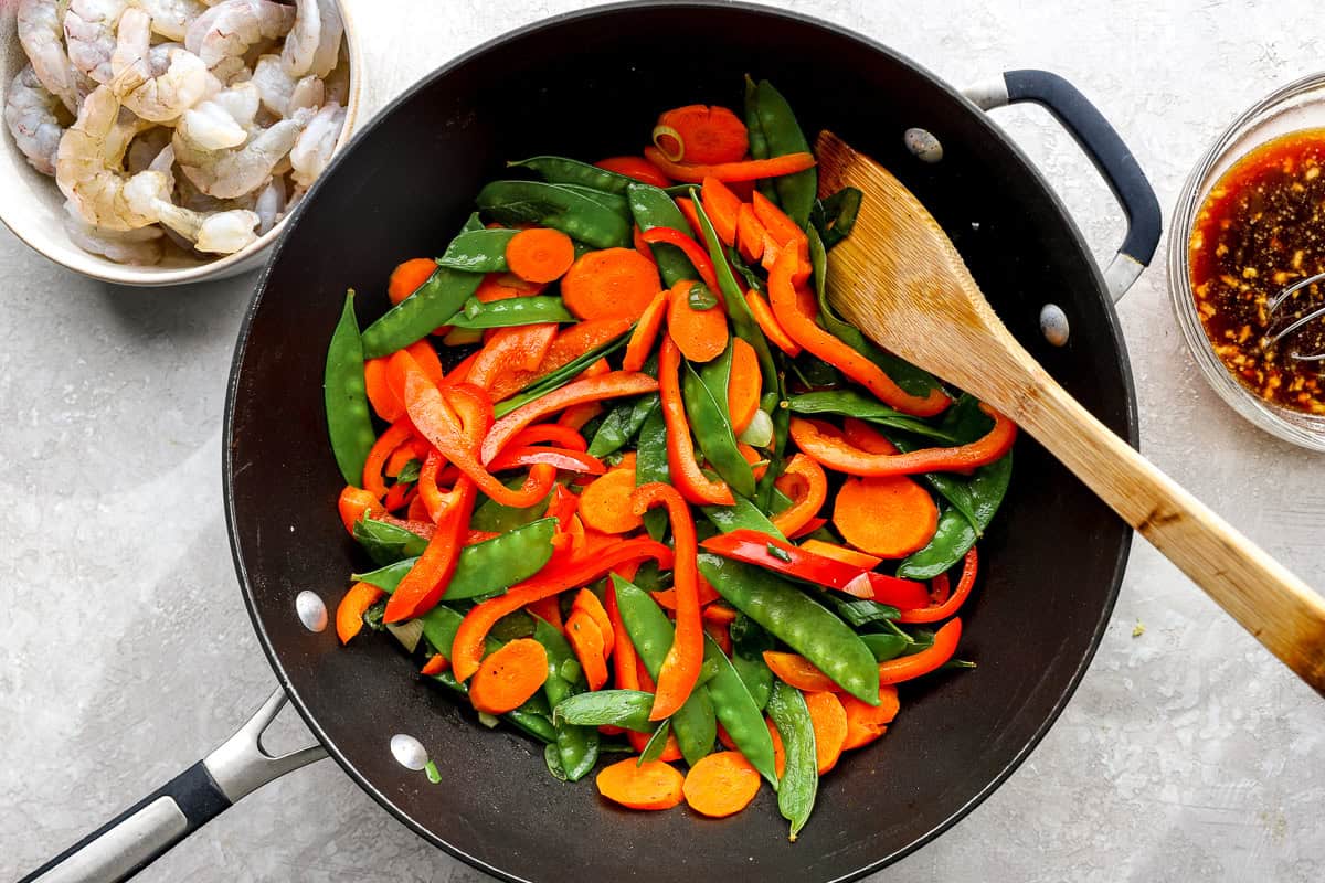 Fresh vegetables including carrots, bell peppers, and snow peas cooking in a skillet