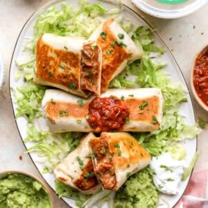 Crispy bean and cheese burritos served with guacamole, salsa, sour cream, and shredded lettuce on a platter.