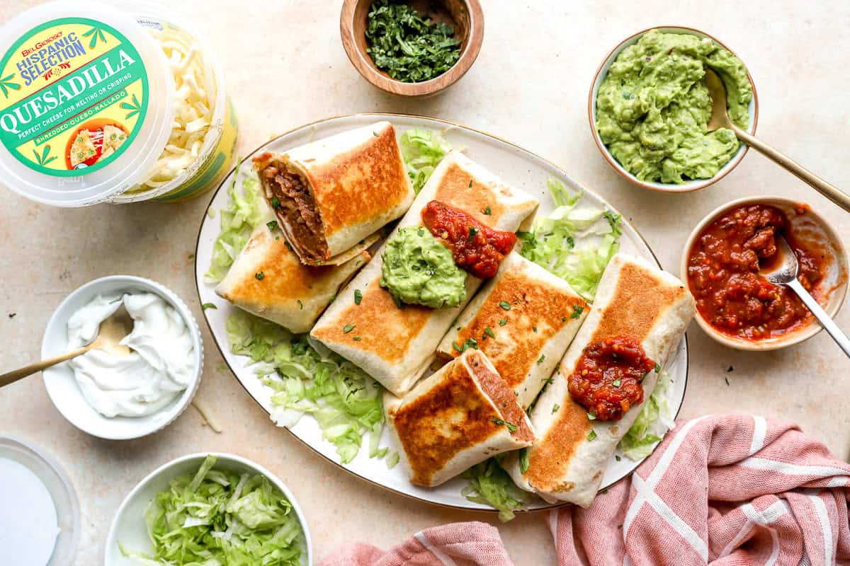 Crispy bean and cheese burritos topped with salsa and guacamole on a serving platter with toppings.