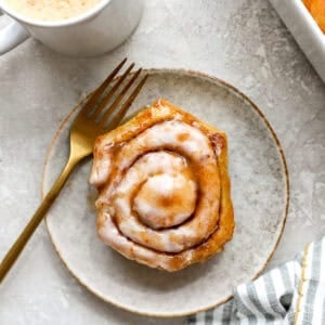 Heavy cream cinnamon roll served on a plate with cream cheese icing and coffee in the background.