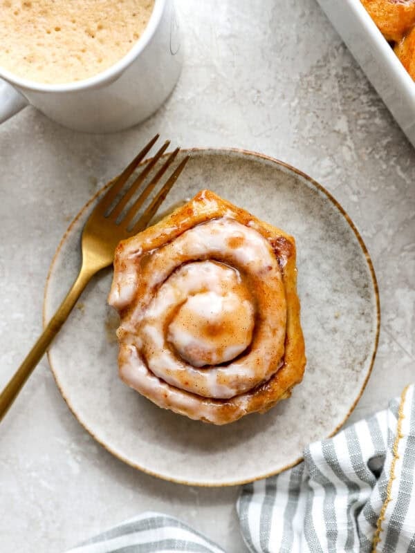 Heavy cream cinnamon roll served on a plate with cream cheese icing and coffee in the background.
