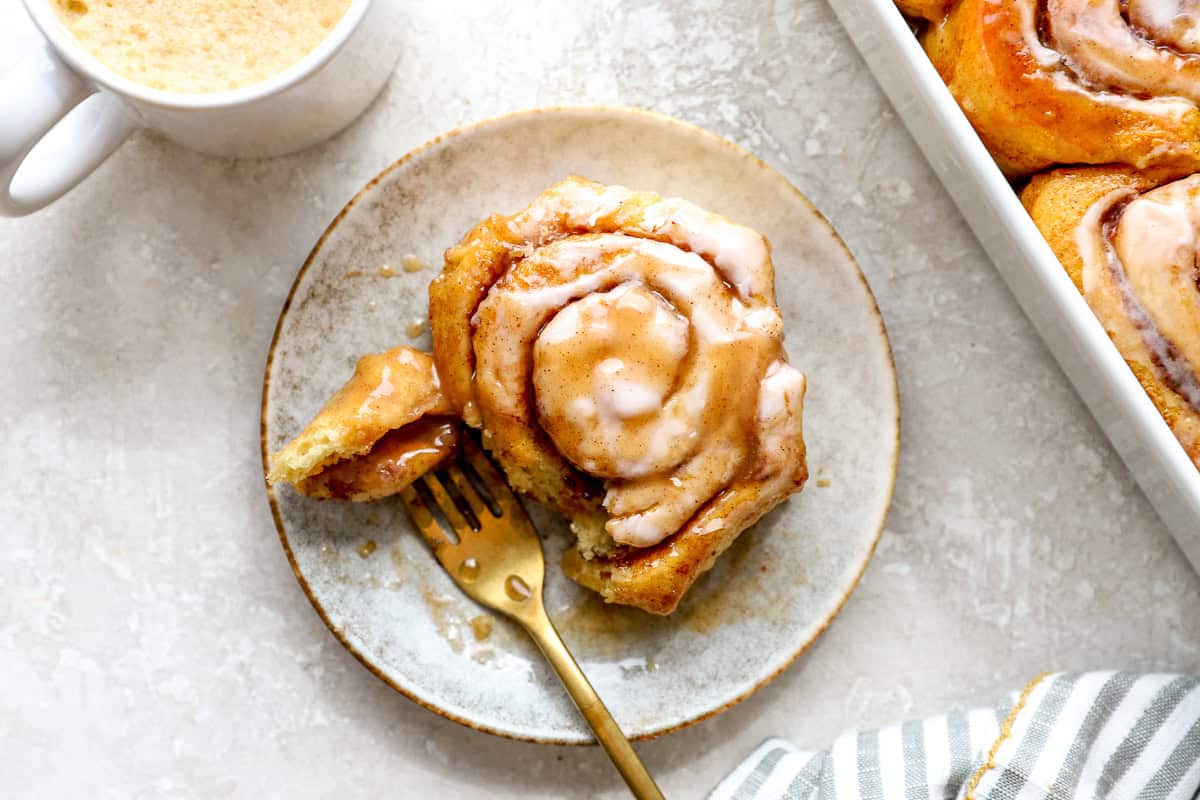 Soft heavy cream cinnamon roll on a plate with a bite removed and coffee in the background.