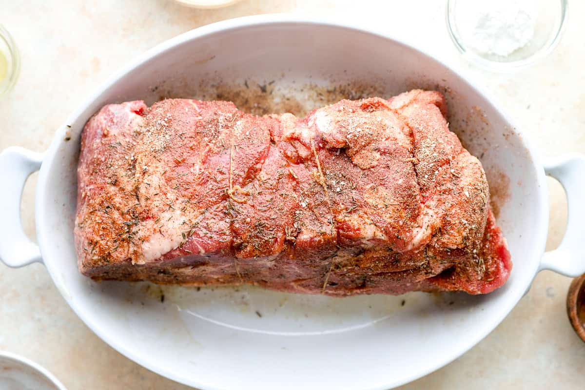 Pork shoulder coated in seasoning blend before roasting in a white baking dish.