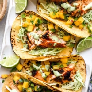 Salmon tacos topped with mango salsa and cilantro lime slaw on charred corn tortillas with fresh lime wedges.