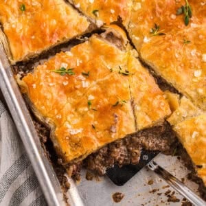 Beef Wellington pot pie bake cut into squares with a spatula lifting a serving from the pan, topped with flaky golden pastry and fresh thyme