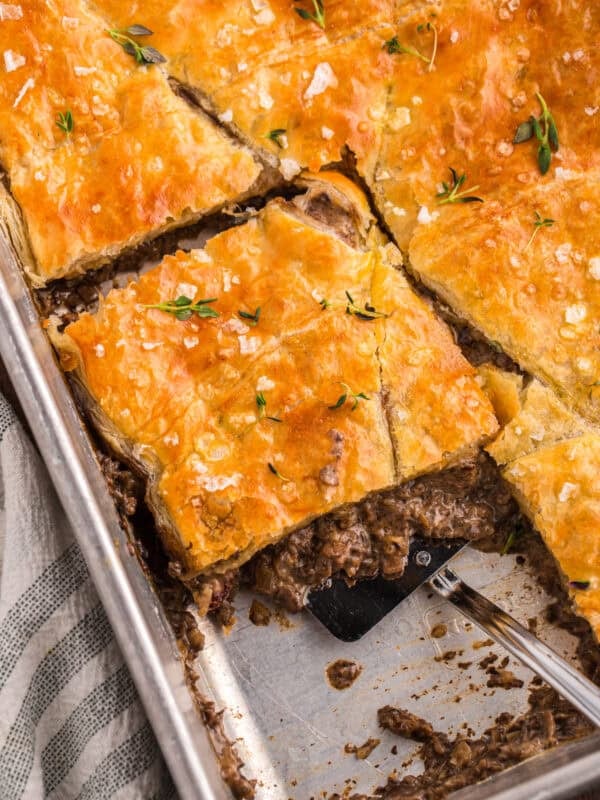 Beef Wellington pot pie bake cut into squares with a spatula lifting a serving from the pan, topped with flaky golden pastry and fresh thyme