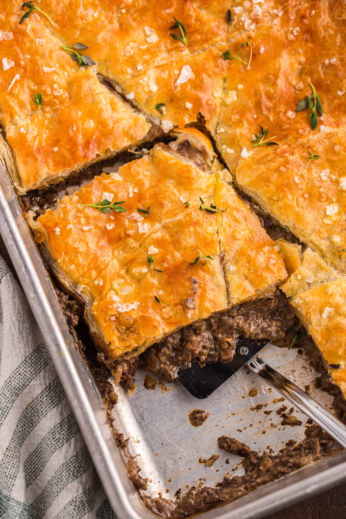 Beef Wellington pot pie bake cut into squares with a spatula lifting a serving from the pan, topped with flaky golden pastry and fresh thyme
