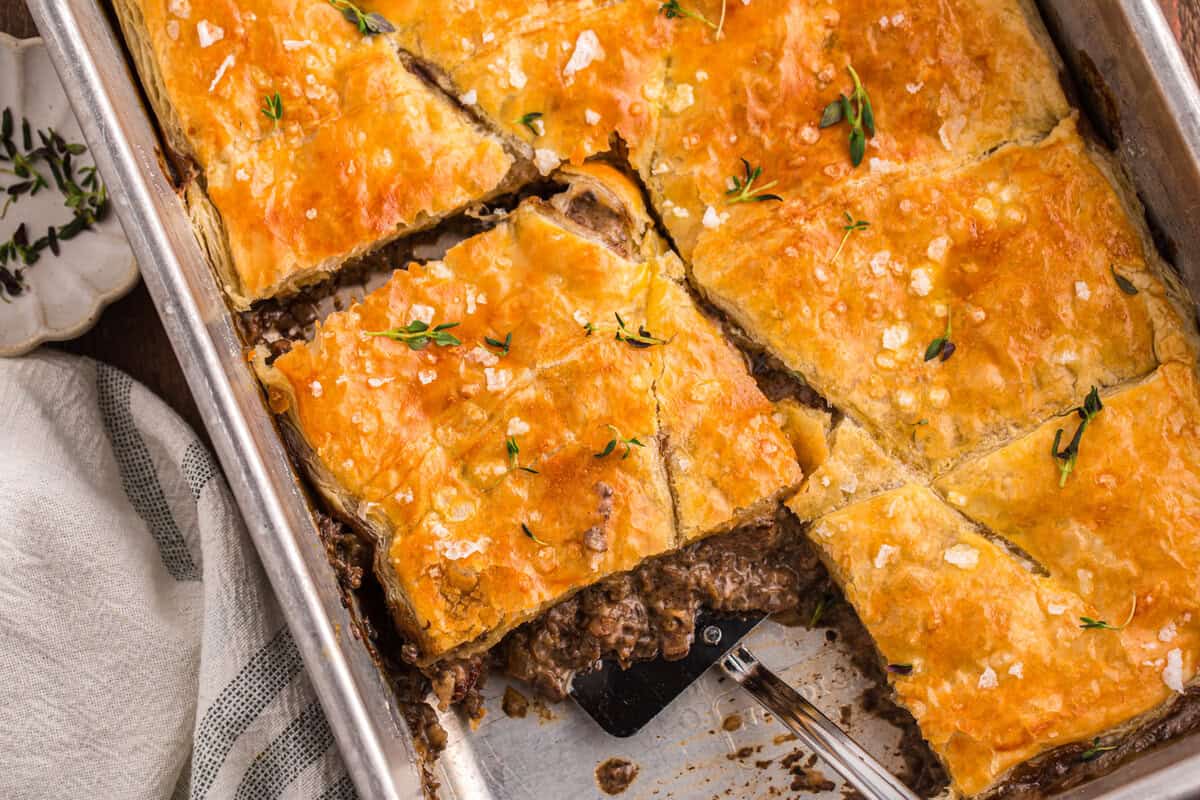 Beef Wellington pot pie bake cut into portions with a spatula lifting a square serving, showing the rich mushroom filling beneath the golden pastry