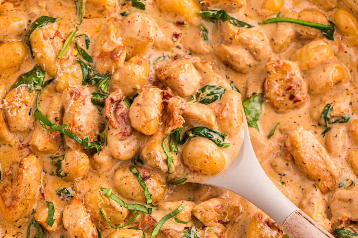 Close-up of marry me chicken gnocchi in a skillet with a spoon scooping pillowy gnocchi, sun-dried tomatoes, and wilted basil in a silky cream sauce.