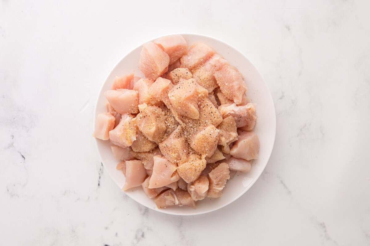 Raw chicken breast cut into bite-sized pieces seasoned with salt, pepper, and garlic powder on a white plate.