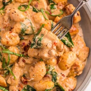 Overhead close-up of marry me chicken gnocchi in a bowl with a fork lifting a piece of chicken, showing the creamy sun-dried tomato sauce and wilted spinach.