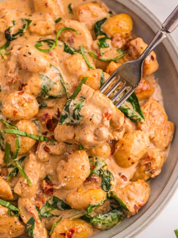 Overhead close-up of marry me chicken gnocchi in a bowl with a fork lifting a piece of chicken, showing the creamy sun-dried tomato sauce and wilted spinach.