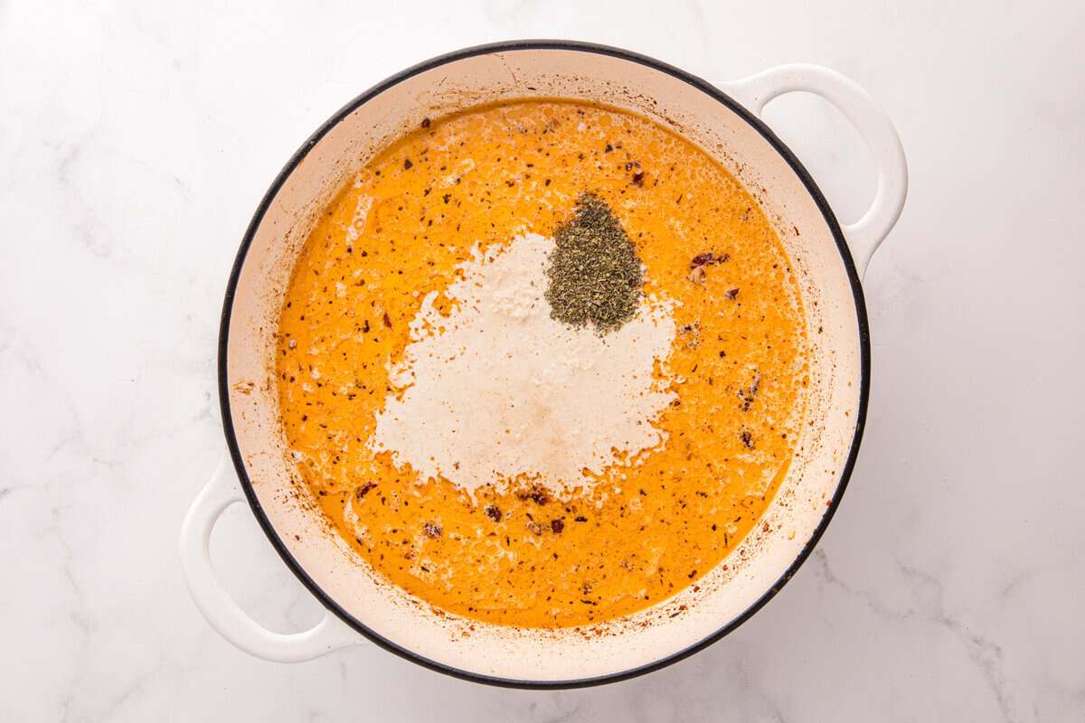 Creamy orange sun-dried tomato sauce in a white skillet with parmesan and Italian seasoning being added.