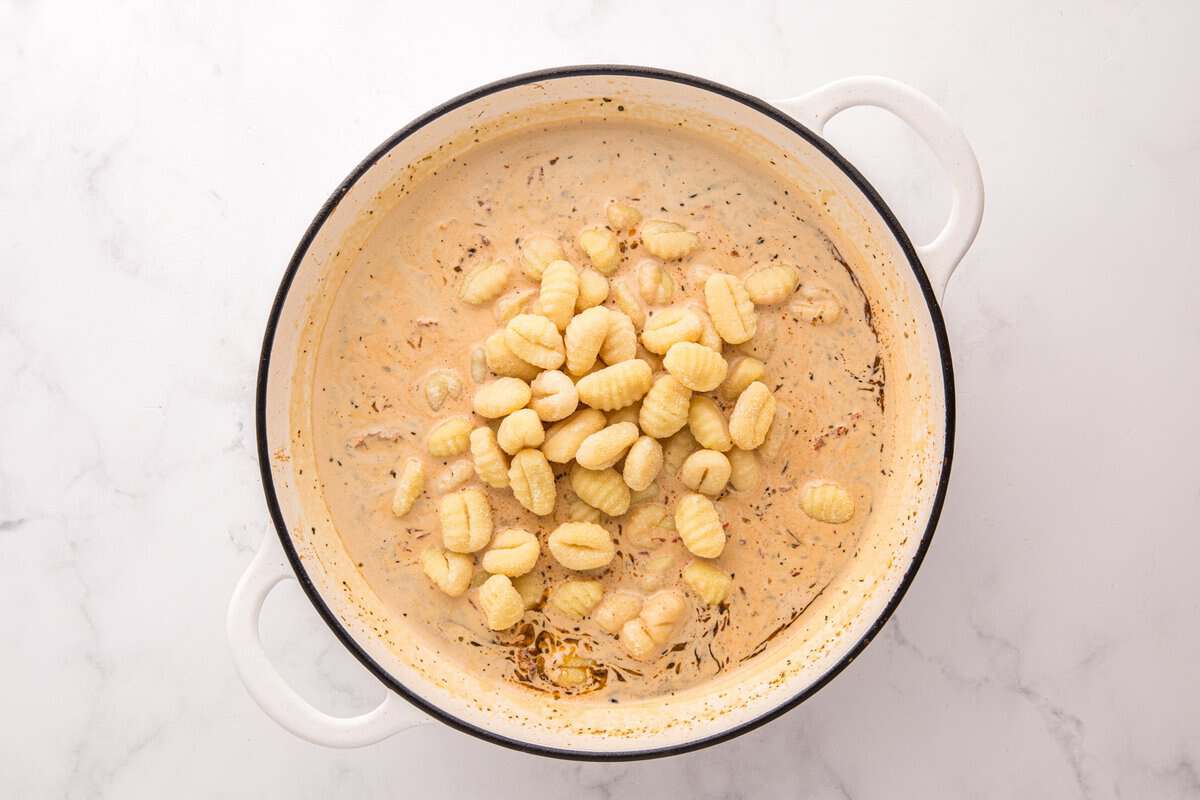 Uncooked potato gnocchi added directly into the creamy sun-dried tomato sauce in a white skillet.